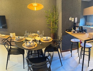 Un comedor con una mesa redonda dispuesta para cuatro personas, rodeada de sillas negras. Cerca de la mesa hay una planta en maceta, y se observa una zona de cocina al fondo.