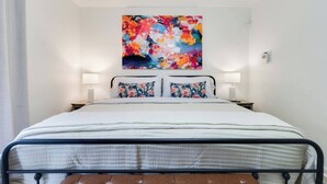 A bed with a metal frame, two pillows with floral patterns, and a colorful abstract painting above it. - Cozy 1BR Condo w/Balcony - Near UL & Ochsner - Apartment (Lafayette)