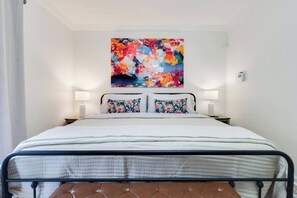 Ein Bett mit Metallgestell, zwei Kissen mit Blumenmuster und ein farbenfrohes abstraktes Gemälde darüber.