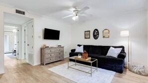 A living room with a black sofa, a wooden cabinet, a coffee table, and a ceiling fan. - Cozy 1BR Condo w/Balcony - Near UL & Ochsner - Apartment (Lafayette)