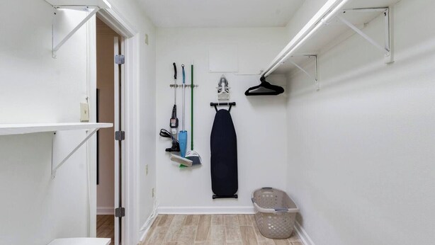 A narrow hallway with a white door, a shelf with cleaning supplies, a hanging coat, and a laundry basket. - Cozy 1BR Condo w/Balcony - Near UL & Ochsner - Apartment (Lafayette)