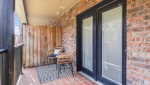 A covered patio area with a brick wall, two sliding glass doors, and a small table with chairs. - Cozy 1BR Condo w/Balcony - Near UL & Ochsner - Apartment (Lafayette)