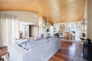 A spacious living area with a large grey sofa, wooden flooring, and a high ceiling with wooden beams.