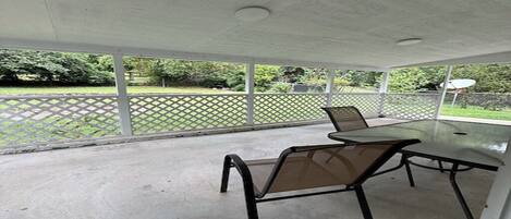 A covered patio area with a table and chairs, a concrete floor, and a view of a green fence and trees.