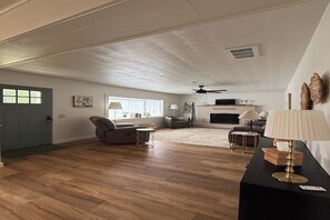 Een ruime woonkamer met houten vloeren, een open haard, een plafondventilator en een deur met een raam.
