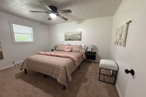 Een slaapkamer met een bed, een plafondventilator, een nachtkastje, een stoel, een raam en een kunstwerk aan de muur.