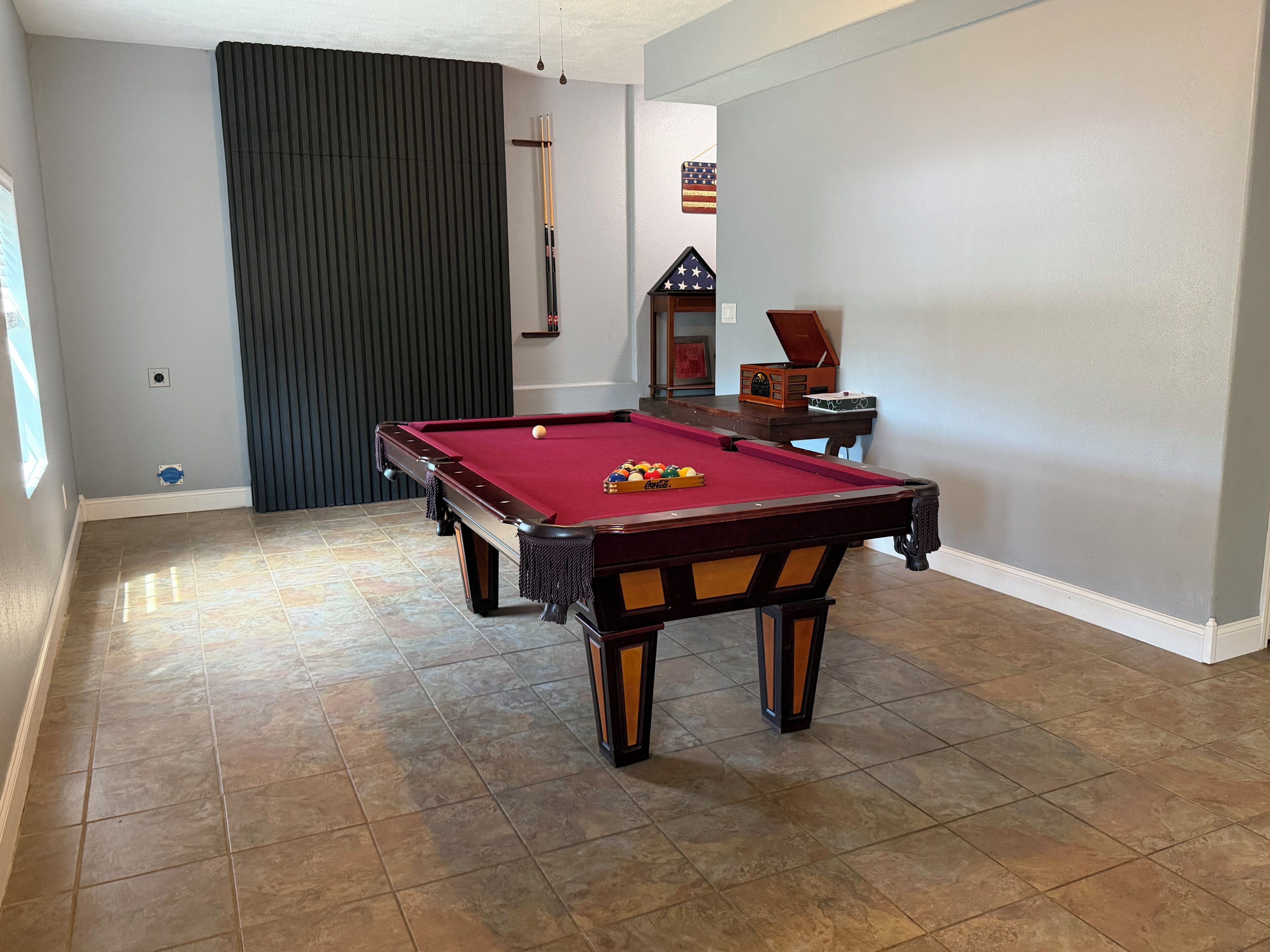 A pool table with a red felt surface, surrounded by a tiled floor, in a room with light-colored walls and a dark-colored partition.