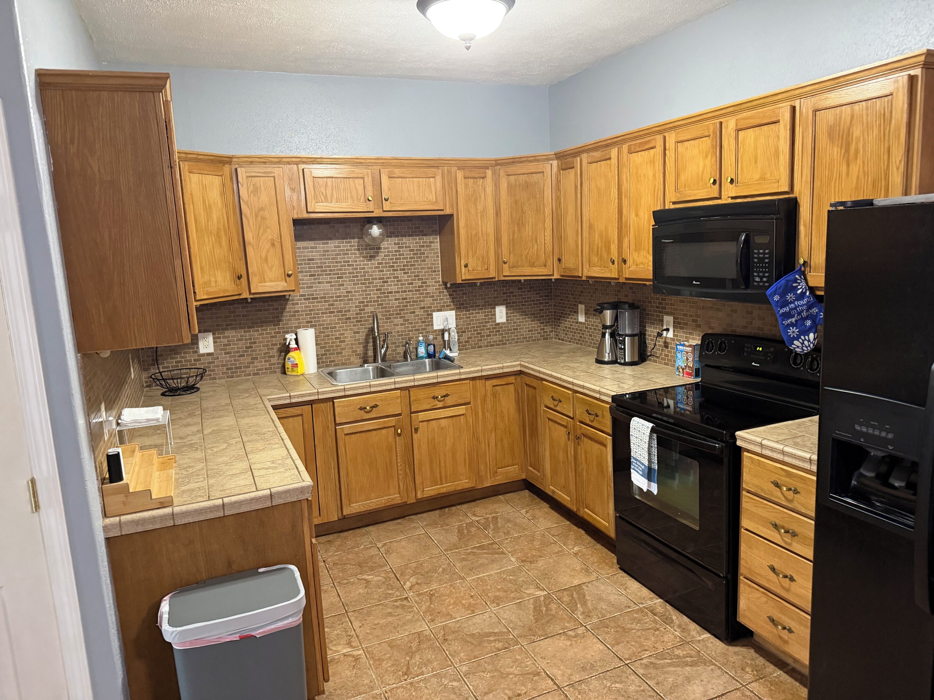 A kitchen with wooden cabinets, a stainless steel sink, a microwave, a stove, and a trash can.