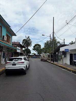 A street with a white car parked on the side, a truck in the distance, and buildings with various signs and awnings. - Cozy 1-bedroom apartment in brilliant Quepos with AC (Quepos)