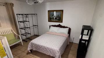 A bedroom with a bed, a standing fan, a framed picture on the wall, and a small table with a box.
