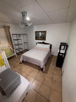 Ein Schlafzimmer mit einem Bett, einem Standventilator, einem gerahmten Bild an der Wand und einem kleinen Tisch mit einer Schachtel.