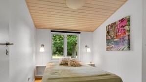 Ett sovrum med en säng, två sänglampor, ett fönster med utsikt över grönområden och en färgstark, abstrakt målning på väggen.