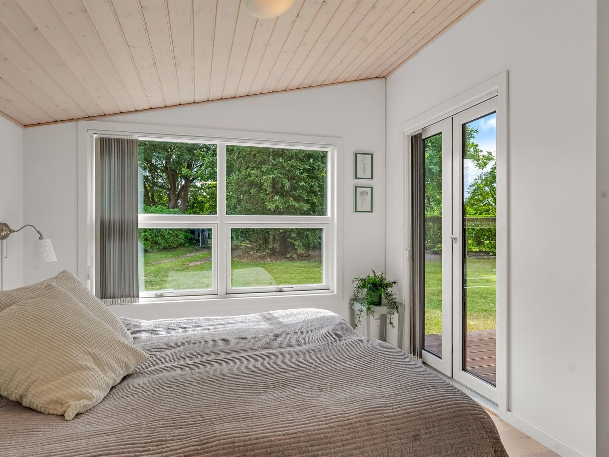 A bedroom with a large window, a bed, a bedside lamp, and a view of a garden.