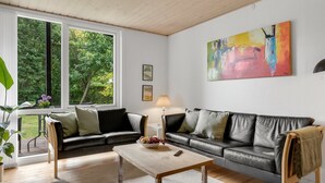 Ett modernt vardagsrum med en svart lädersoffa, ett träbord och ett stort fönster med utsikt över grönområden.