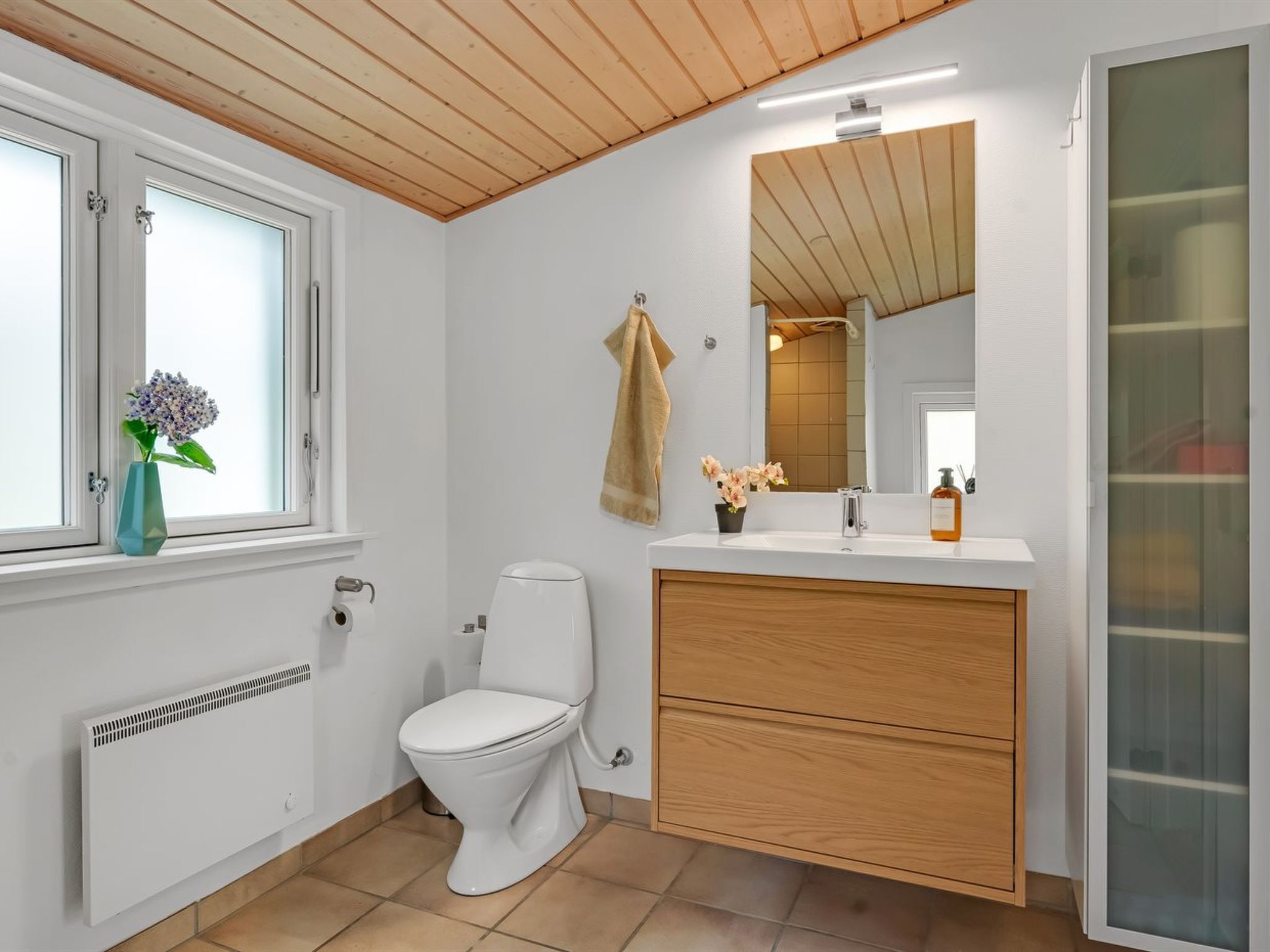 A bathroom with a toilet, a sink with a mirror, a towel hanging on the wall, and a window with a vase of flowers.