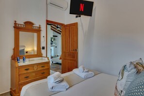 Ein Hotelzimmer mit einem hölzernen Schminktisch, einem Bett mit Handtüchern, einem an der Wand montierten Fernseher und einer Tür zu einem weiteren Raum.