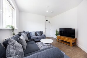 Ein modernes Wohnzimmer mit einer Eckcouch, einem Flachbildfernseher auf einem Holzgestell und einem kleinen weißen Tisch.