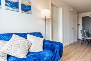 A room with a blue textured sofa, a floor lamp, and a dining area with chairs and a table. - Modern 1-Bed City Centre Apartment in Digbeth – Sleeps 2 (Birmingham)