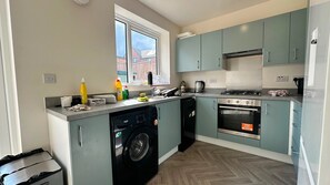 A compact kitchen with grey cabinets, a dishwasher, a stove, and a window with a view of a house. - Sheffield Charm - Cozy 2 Bedroom House (Sheffield)