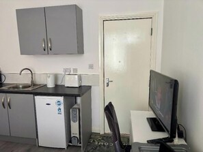 Eine kompakte Küchenzeile mit einem Kühlschrank, einer Kaffeemaschine und einer Mikrowelle. Eine Tür mit Griff und ein kleines Fenster sind vorhanden.