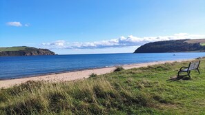 A bench on a grassy area overlooking a beach with a calm sea and distant hills.