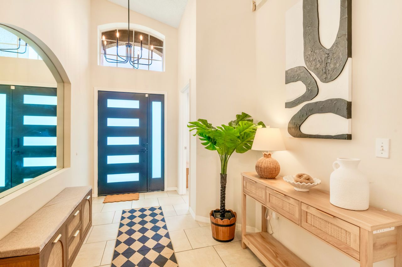 A hallway with a checkered rug, a potted plant, a wall-mounted abstract art piece, a wooden console table with decorative items, and a door with horizontal slats.