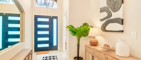 A hallway with a checkered rug, a potted plant, a wall-mounted abstract art piece, a wooden console table with decorative items, and a door with horizontal slats.