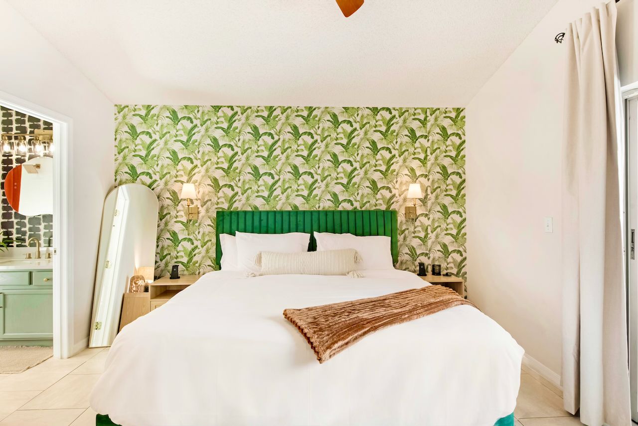 A hotel room with a bed, green headboard, two bedside lamps, and a window with curtains.
