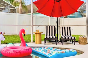 Ein Poolbereich mit einem roten Sonnenschirm, zwei gestreiften Liegestühlen und einem Flamingo-Schwimmpflege.