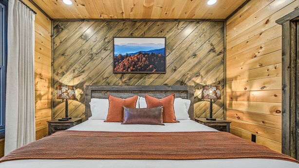 A bedroom with a wooden wall, a bed with pillows, a nightstand, and a framed picture on the wall. - Happy Chipmunk - Mountain Views, Hot Tub, Fire Pit & Game Room (Sevierville)