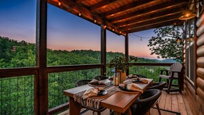 A wooden deck with a table set for dining, overlooking a forested landscape. - Happy Chipmunk - Mountain Views, Hot Tub, Fire Pit & Game Room (Sevierville)