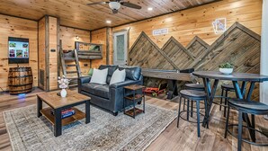 A cozy room with a wooden ceiling, a ceiling fan, a flat-screen TV, a leather sofa, a coffee table, a small table with a plant, and a bar area with stools. - Happy Chipmunk - Mountain Views, Hot Tub, Fire Pit & Game Room (Sevierville)