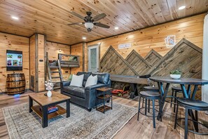 A cozy room with a wooden ceiling, a ceiling fan, a flat-screen TV, a leather sofa, a coffee table, a small table with a plant, and a bar area with stools. - Happy Chipmunk - Mountain Views, Hot Tub, Fire Pit & Game Room (Sevierville)