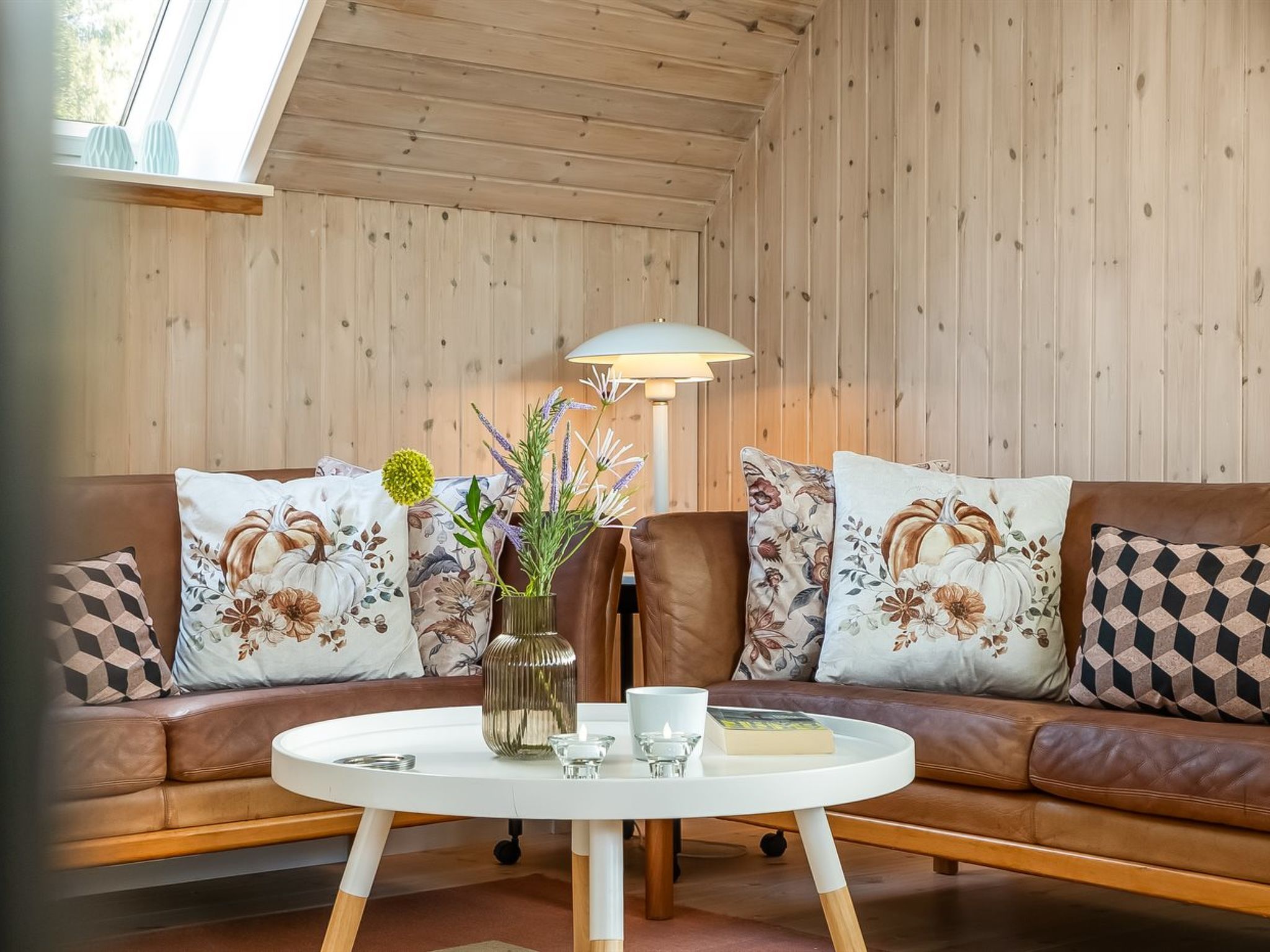 A cozy living room with a brown leather sofa, floral cushions, a round white coffee table, and a floor lamp.