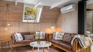 L’intérieur chaleureux d’un chalet avec un revêtement mural en bois, une grande fenêtre, un canapé en cuir marron, une table basse blanche et un appareil de climatisation.