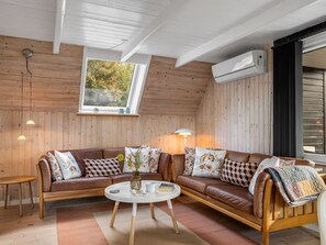 Ein gemütliches Blockhaus-Interieur mit Holzvertäfelung, einem großen Fenster, einem braunen Ledersofa, einem weißen Couchtisch und einer Klimaanlage.