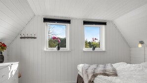 Une chambre à coucher avec un lit, une commode, deux fenêtres ornées de plantes et un plafond en pente.