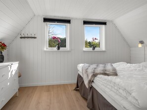 Ein Schlafzimmer mit einem Bett, einer Kommode, zwei Fenstern mit Pflanzen und einer geneigten Decke.