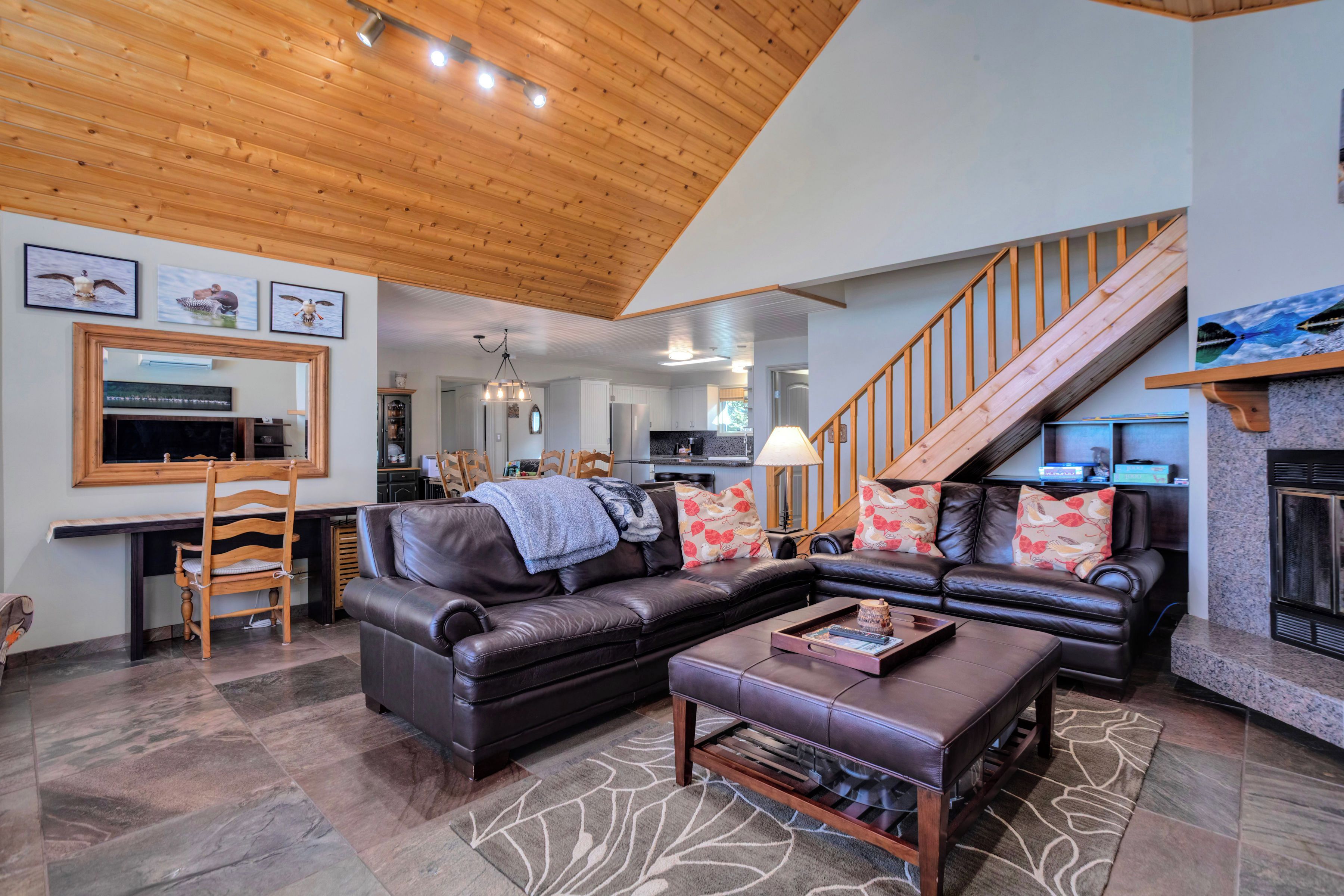 A spacious living room with a wooden ceiling, a black leather sofa, a coffee table, and a staircase leading to an upper level.