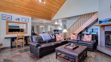 A spacious living room with a wooden ceiling, a black leather sofa, a coffee table, and a staircase leading to an upper level.