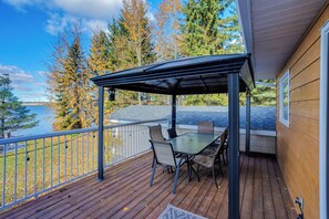 A covered outdoor deck with a table and chairs, overlooking a body of water and trees.