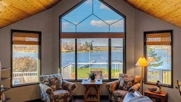 A cozy room with a large window offering a view of a lake and trees. There are two patterned armchairs and a small table with a lamp.
