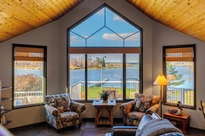 A cozy room with a large window offering a view of a lake and trees. There are two patterned armchairs and a small table with a lamp.