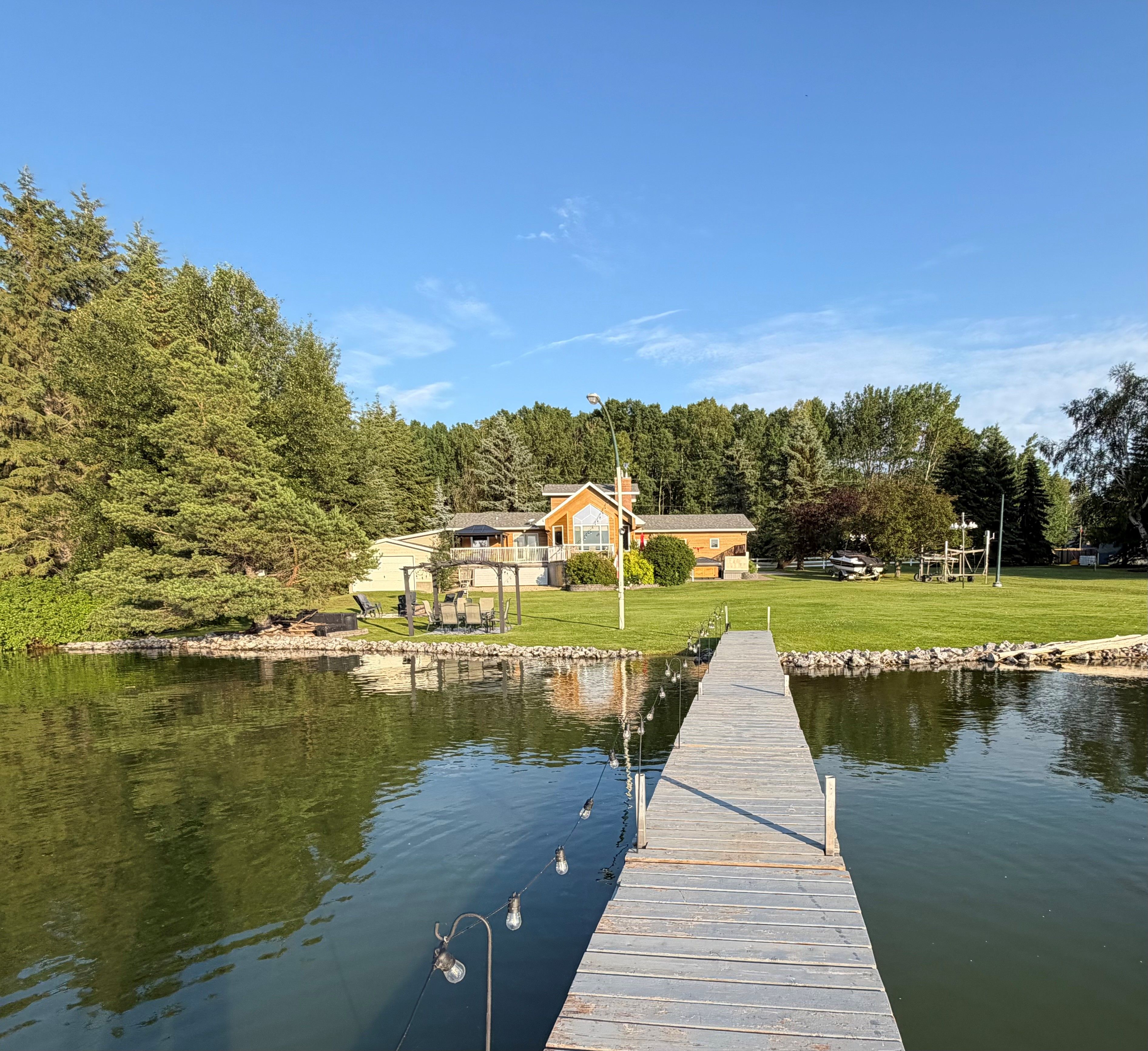 A wooden dock extending into a calm body of water, surrounded by a grassy area with trees and a building in the background.
