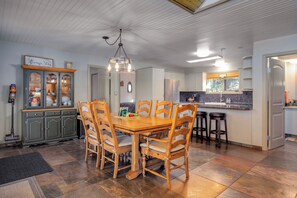 A dining area with a wooden table and chairs, a chandelier, and a kitchen in the background.