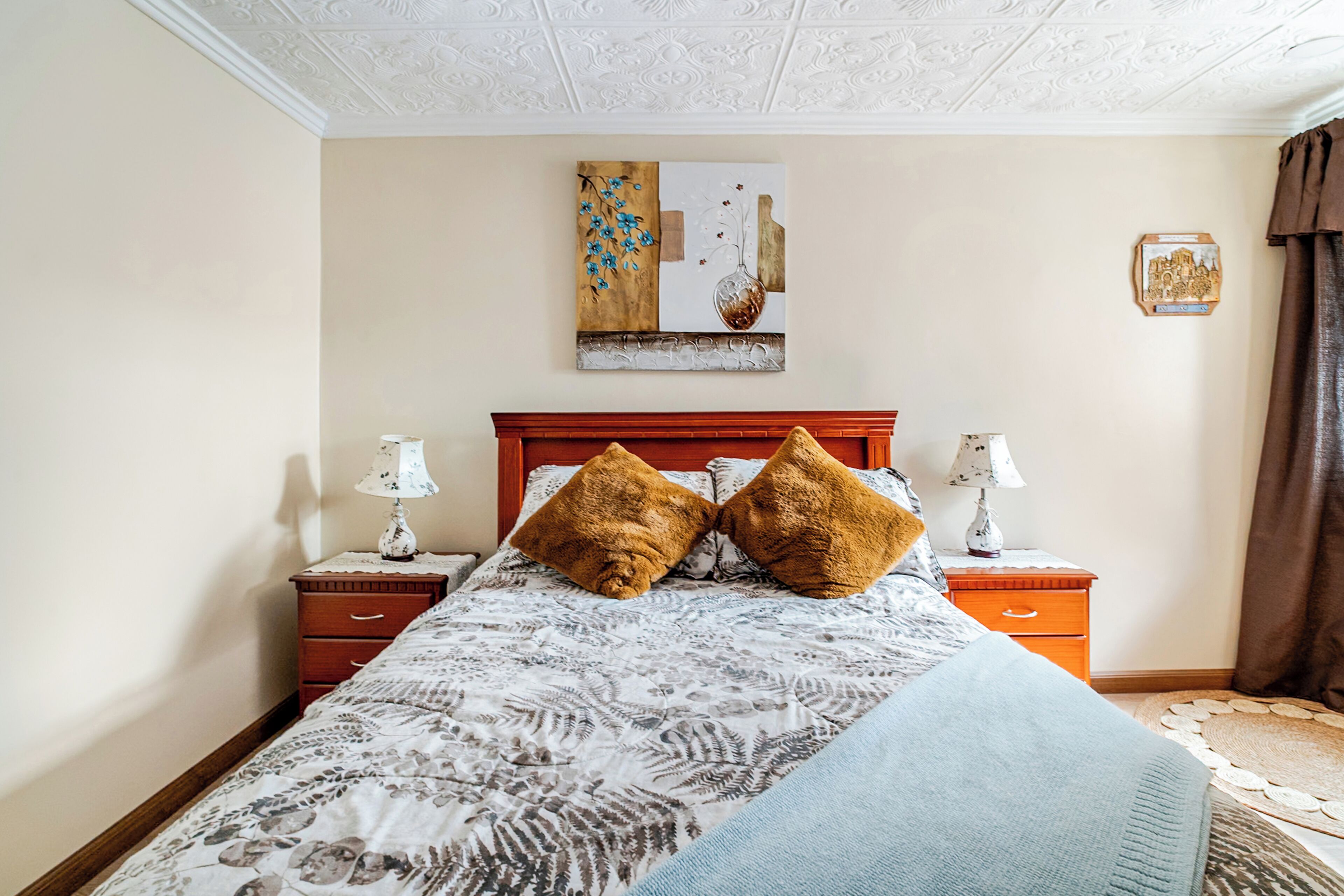 Un dormitorio con una cama, dos mesitas de noche, un cabecero de madera, un adorno en la pared y una lámpara de noche a cada lado.