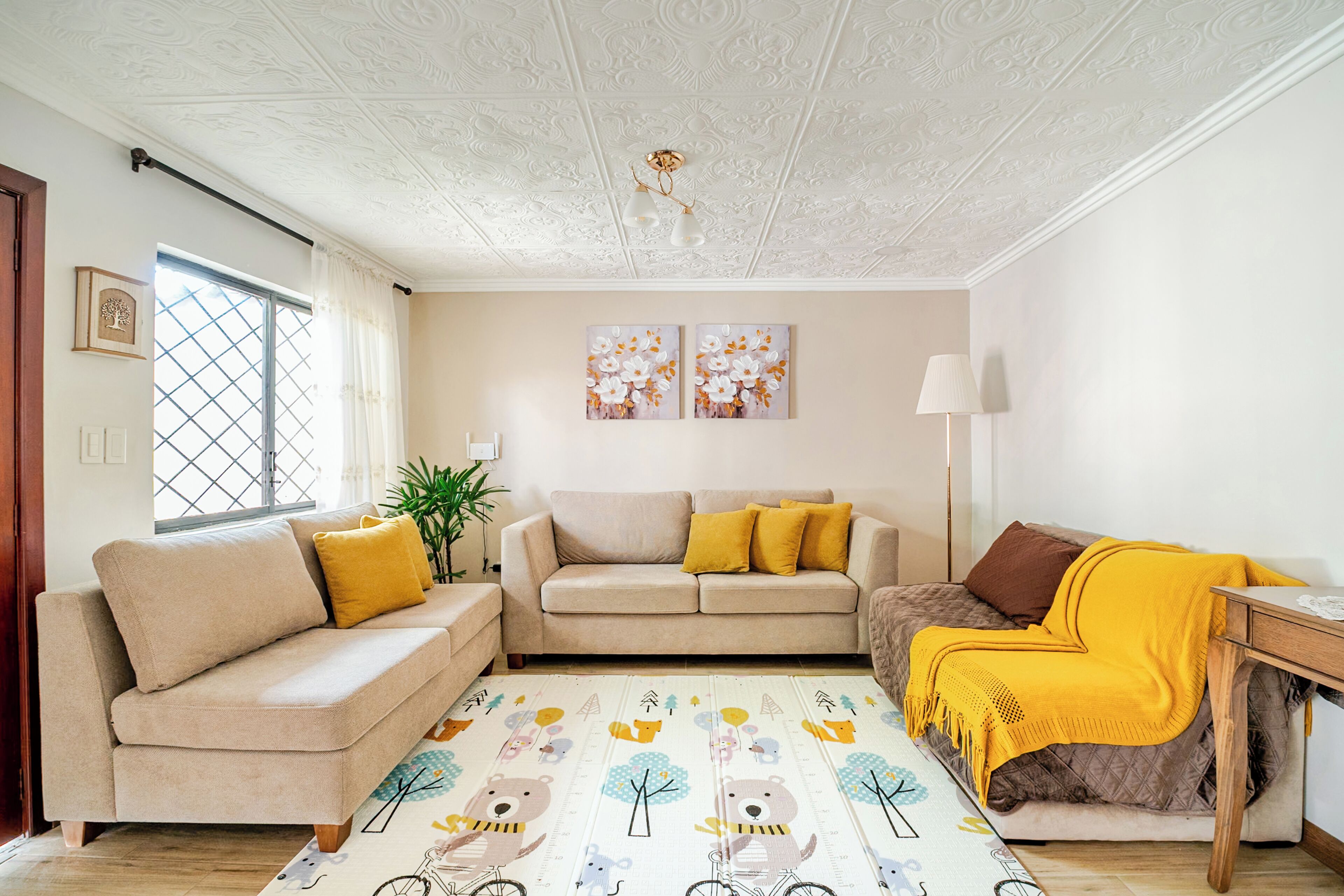 Un salón con un conjunto de sofá, una mesa de centro y una mesita auxiliar con una lámpara. Hay una ventana con cortinas, una planta y dos cuadros colgados en la pared.