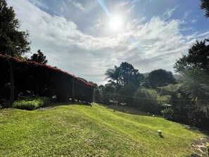 A well-maintained grassy area with a variety of trees and shrubs, a clear sky, and a sun shining through the clouds. - Hekerua Bay Views (Waiheke Island)