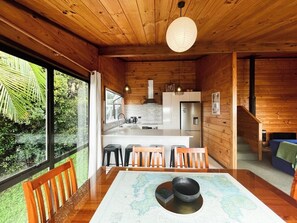 A wooden interior with a dining table, chairs, a kitchen area, and a view of greenery outside. - Hekerua Bay Views (Waiheke Island)