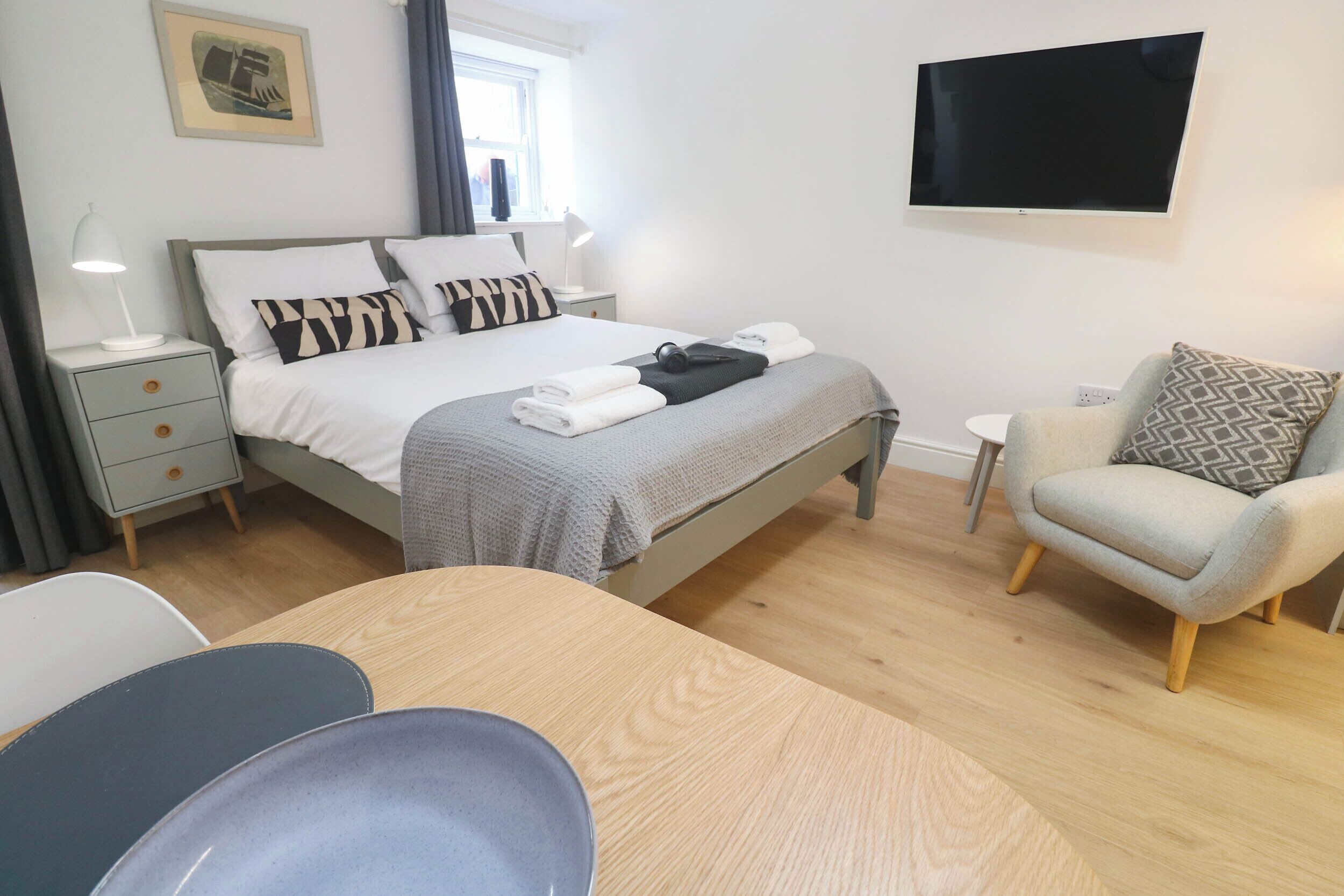 A hotel room with a bed, bedside table, lamp, chair, and a flat-screen TV mounted on the wall.